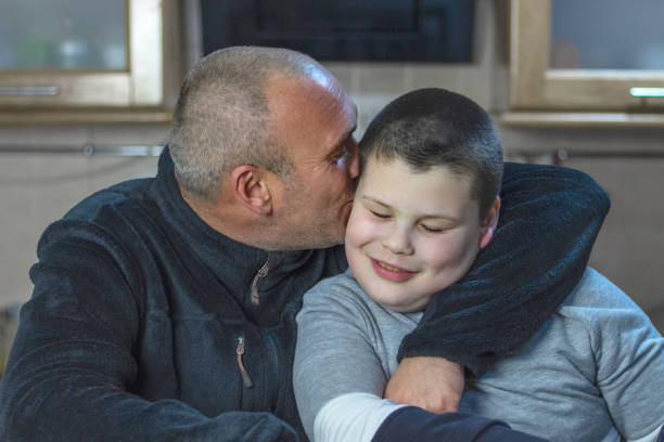 Dad and his son with autism sitting in kitchen at home. A man with short hair and bristle hugs his son and kisses him on the cheek.  autism stock pictures, royalty-free photos & images