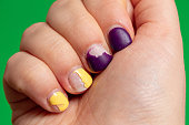 istock Damaged nail polish of purple and yellow color on the nails of the hand. Green background with space for text. Close-up. Selective focus. 1319928756