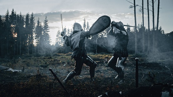 Dark Age Battlefield: Two Armored Medieval Knights Fighting with Swords. Battle of Armed Warrior Soldiers, Killing Enemy in Mysterious Forest. Cinematic Smoke, Mist, Light in Historic Reenactment