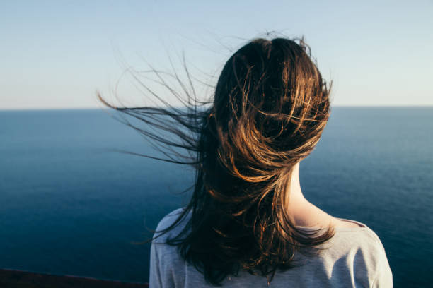 Dark hair girl's portrait. Woman with dark hair stands on a top cliff over blue sea view while wind. rear view stock pictures, royalty-free photos & images