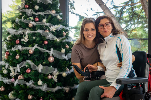 A daughter and mother with a disability - with dystrophy  in a wheelchair decorates a Christmas tree in the hotel’s large lobby with beautiful views of the green treetops in a seaside Mediterranean town. stock photo
