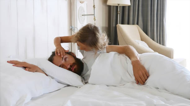 Daughter comes to sleeping father and opens his eyes by hands for wakes him Little cute daughter comes to sleeping father at morning, and opens his eyes by fingers for wakes him. Family at home concept. waking up stock pictures, royalty-free photos & images