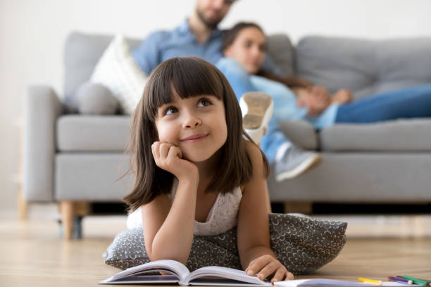 Daughter dreaming lying on warm floor with book at home Close up little adorable thoughtful smiling daughter dreaming lying at cushion on warm floor with book in living room at modern home, resting married couple parents on background, focus on small kid comfortable stock pictures, royalty-free photos & images