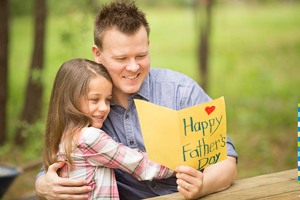 Daughter shows dad handmade Father's Day card. Outdoors. Child, parent. Mid-adult caucasian father excitedly receives a homemade card from his daughter. The cute paper card reads "Happy Father's Day."  They give each other a big hug.  This parent and child are sitting at an outdoor table in their backyard. Beautiful summer or spring nature background. fathers day stock pictures, royalty-free photos & images