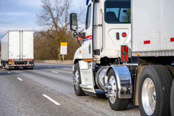 Day cab white big rig semi truck with chrome fenders transporting cargo in dry van semi trailer running in convoy behind another semi truck driving on the interstate highway road stock photo