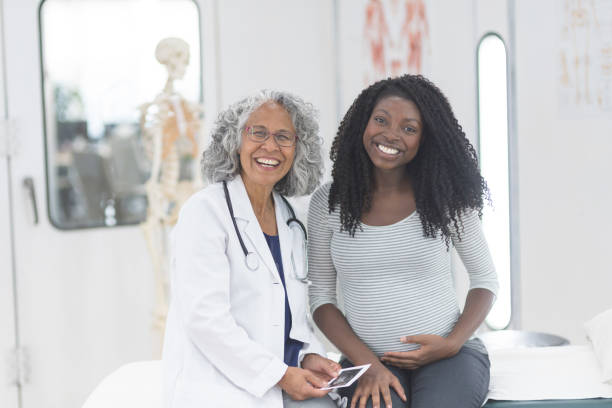 Day in the Life of a Patient An ethnic female OB/GYN doctor sits on an examination table next to her pregnant patient. They are looking at an ultrasound picture and are both smiling at the camera. obstetrician photos stock pictures, royalty-free photos & images
