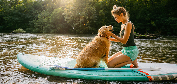 Photo of a smiling young woman and her dog enjoying the beautiful, warm summer afternoon by the river, far from the hustle of the city.