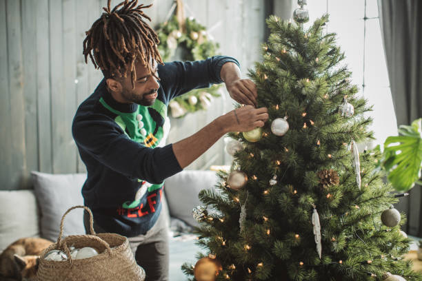 Decorating a Christmas tree Young man celebrating Christmas at home and decorating christmas tree. Home is decorated with Christmas ornaments and lights. chrismas tree stock pictures, royalty-free photos & images