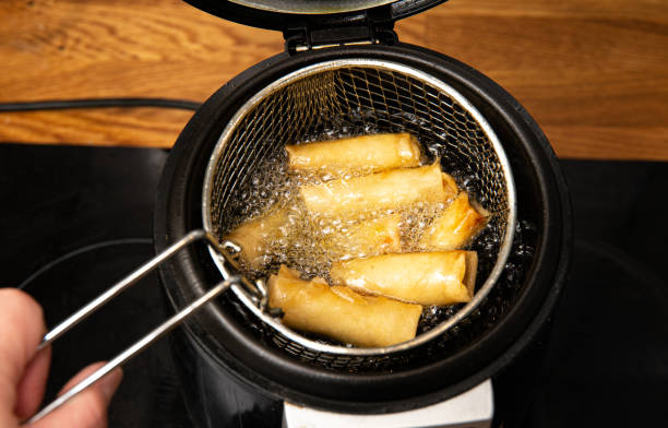 Deep fried fritted vegetable spring rolls inside special deep fryer electrical machine pot, boiling in hot cooking oil. Crispy golden look. hand lifting up sieve. stock photo
