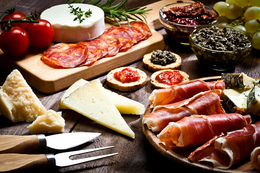 Rustic wood table filled with a assortment of fine food. A wood tray with prosciutto and cheese is at the bottom-right of the frame. Two glass bowls with dried tomatoes and olives paste and a cutting board are at the top. Various types of cheese complete de composition. DSRL studio photo taken with Canon EOS 5D Mk II and Canon EF 100mm f/2.8L Macro IS USM