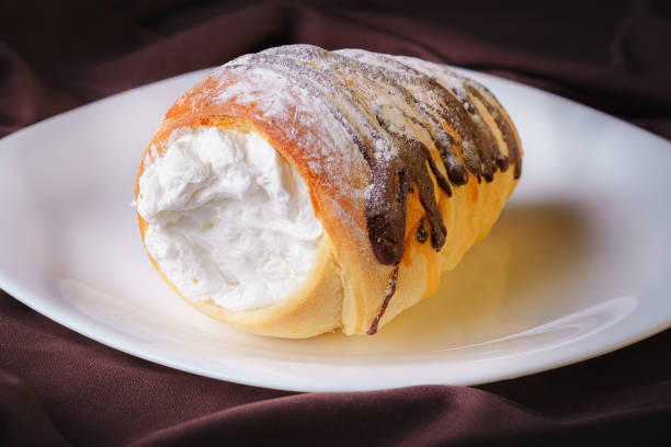 delicious rolled cone shaped bread filled with pastry cream covered with homemade chocolate and sugar placed on a white ceramic plate stock photo