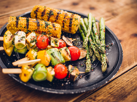 High angle view of a healthy selection of grilled vegetable kebabs, asparagusand corn on a dark plate on a wooden surface