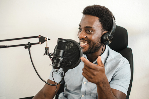 Glad African American male radio host in headphones sitting at table and speaking in microphone while recording podcast in studio on white background