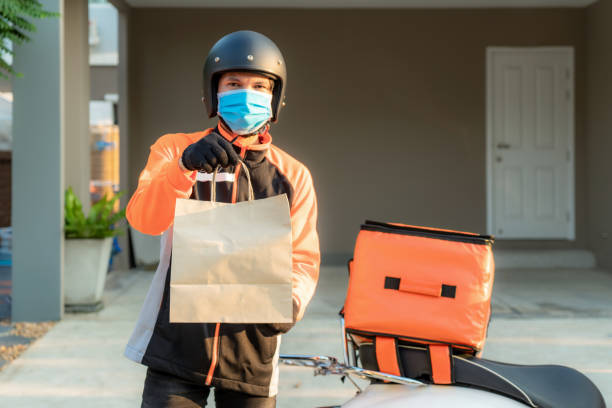 배달 아시아 남자는 오렌지 유니폼에 보호 마스크를 착용하고 스쿠터에 케이스 상자와 고객 집 앞에 배달 음식 가방을 보낼 준비가, 음식 배달 및 쇼핑 온라인 개념. - 배달 뉴스 사진 이미지