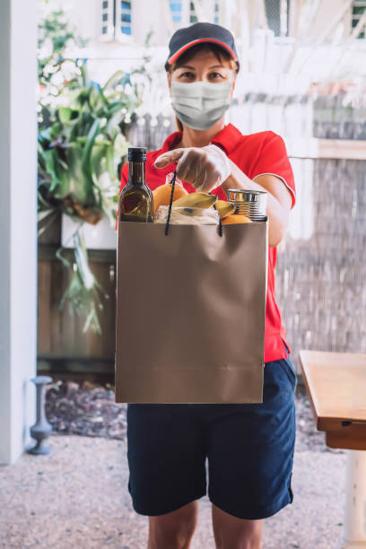 Delivery woman in protective face mask and gloves delivers paper bag with food products. stock photo