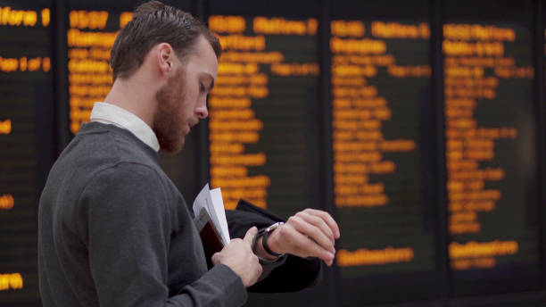 A young beadrded man checking his documents as he stands in front of an out-of-focus electronic departures board (train or airport).