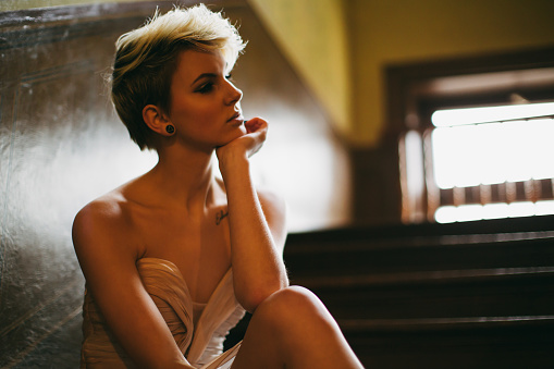 Depressed and angry blonde young woman waering an evening gown sitting on wooden stairway steps of old mansion. Woman Angry - Sadness - Depression Concept.