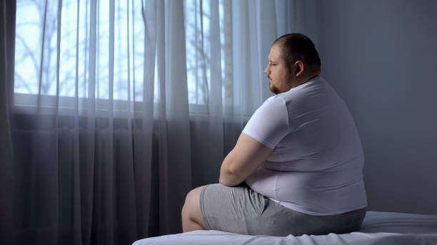 Depressed fat man sitting on bed at home, worried about overweight, insecurities stock photo