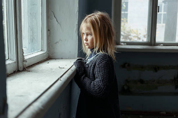 Depressed poot girl standing near window Hopeless life. Close up of depressed poor little girl standing near window and looking aside while feeling miserable poverty stock pictures, royalty-free photos & images