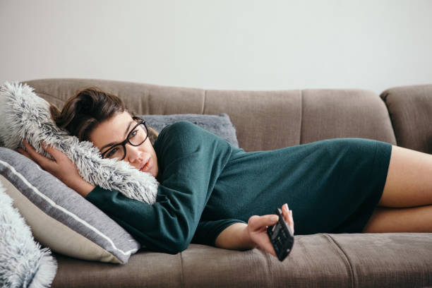 Depressed Woman Changing Channels on a TV Remote Young woman lying down on a sofa, feeling depressed and changing channels on a TV remote. boredom stock pictures, royalty-free photos & images