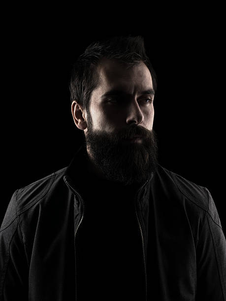 Desaturated serious bearded hipster looking away. Desaturated serious bearded hipster looking away. High contrast low key dark shadow portrait isolated over black background. chiaroscuro stock pictures, royalty-free photos & images