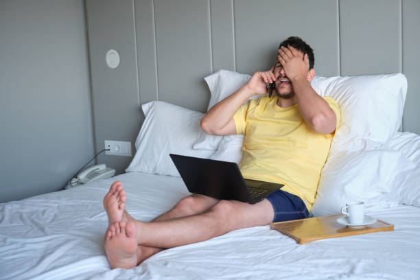 Desperate man speaks on the phone and works with laptop in an hotel room bed. stock photo