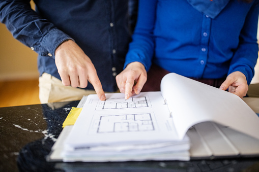 Midsection of couple pointing at floor plan on file. Man and woman are analyzing blueprint. They are standing at kitchen island.