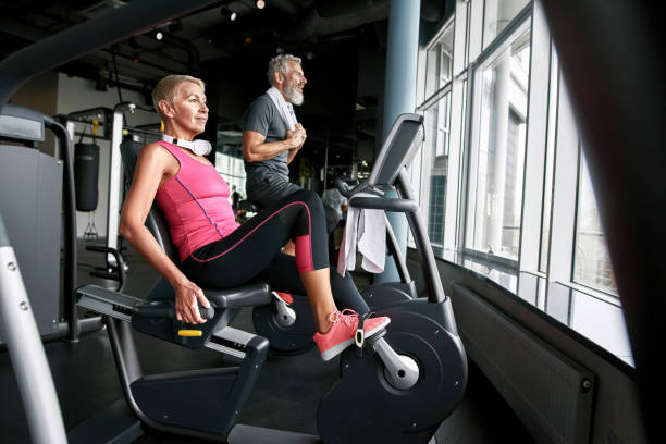 Determined senior couple working out on excercise bikes. Beautiful young looking senior woman in pink top cycling in gym together with elder bearded man in gray t-shirt. Senior couple of determined husband and wife continuing active life after retirement. stationary bike stock pictures, royalty-free photos & images