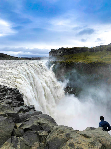 Dettifoss, North Iceland: Adventurous Tourist At Edge of Waterfall Dettifoss, North Iceland: Adventurous Tourist At Edge of Waterfall. Copy space in the sky. dettifoss waterfall stock pictures, royalty-free photos & images