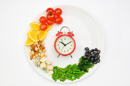 Diet and lunchtime, Intermittent fasting concept. Vegetables, oranges, cheese, nuts and clock on a white plate. Healthy dietary food.
