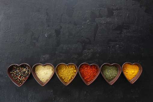 Different kind of spices on a black stone. Oriental spices in in heart shaped bowl, red peppers, curry powder, cinnamon powder, mint powder, colorful peppers. Flat lay, top view.