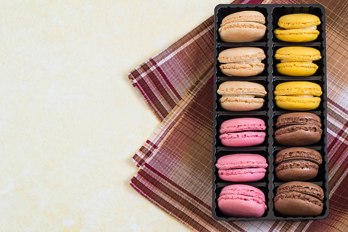 Different macaroon in box on light yellow background with brown checkered napkin. Delicious and sweet almond cookies macaroon with cream, French delicacy