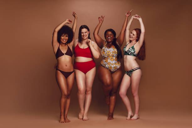 Different size women in bikinis dancing together Full length of different size women in bikinis dancing together over brown background. Multi-ethnic women in swimwear enjoying themselves. body shape woman stock pictures, royalty-free photos & images