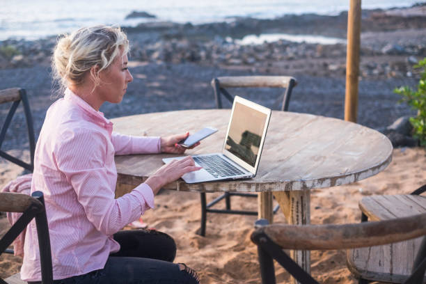 digital nomad concept with blonde free beautiful caucasian middle age woman working at the laptop using cellular phone too sitting on a wood table near the ocean - freelance free to work everywhere with technology digital nomad concept with blonde free beautiful caucasian middle age woman working at the laptop using cellular phone too sitting on a wood table near the ocean - freelance free to work everywhere with technology nomadic people stock pictures, royalty-free photos & images
