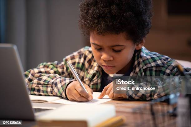 Diligent African schoolboy in casualwear making notes in copybook by table