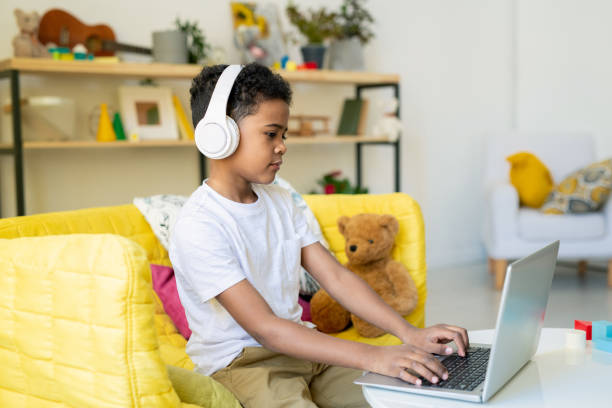 Diligent schoolboy of elementary age in headphones pushing laptop buttons Diligent schoolboy of elementary age in headphones pushing buttons of laptop keypad while studying by table in home environment remote control stock pictures, royalty-free photos & images