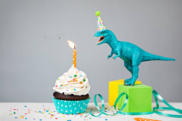 Dinosaur Birthday Party Toy Dinosaur blowing out a Birthday candle with cup cake on a gray background humorous happy birthday images stock pictures, royalty-free photos & images