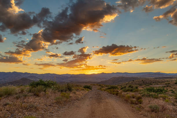 A dirt road leading to distant mountains where the sun has just set behind with colorful clouds stock photo