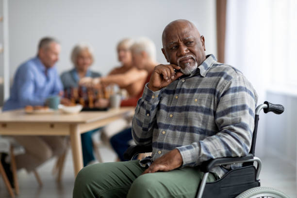 Disabled african american senior man in wheelchair feeling lonely Disabled african american senior man in wheelchair feeling lonely, sitting aside of cheerful elderly men and women playing chess and chatting. Adaptation at retirement house concept sad old black man stock pictures, royalty-free photos & images
