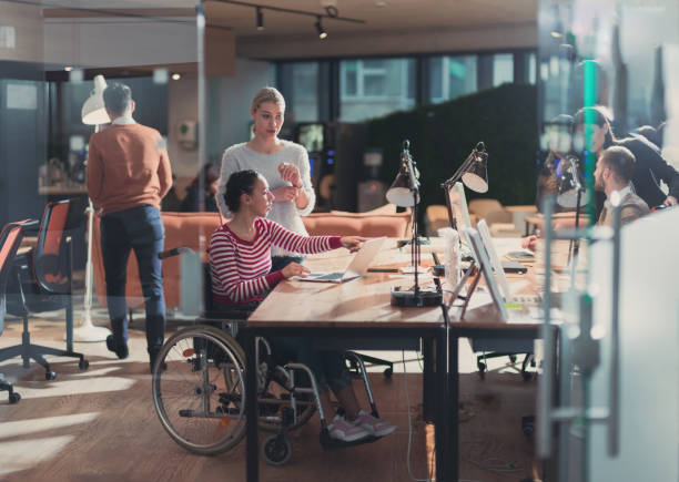 Disabled businesswoman in a wheelchair in modern coworking office space. Colleagues in background. Disability and handicap concept. Selective focus stock photo