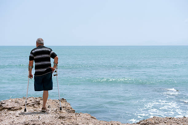 Disabled man on crutches on rocks at the sea stock photo