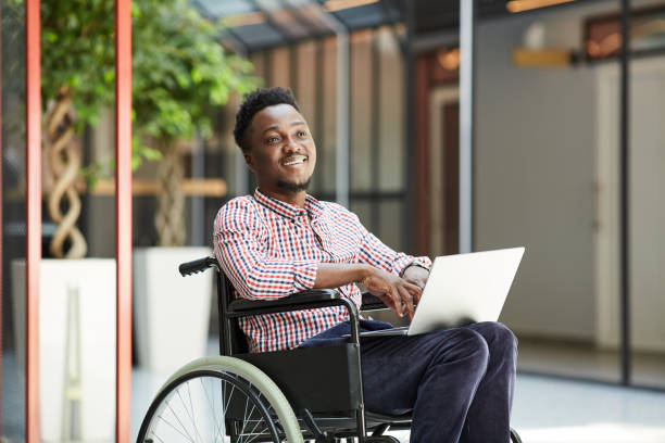 Disabled man using laptop African smiling man sitting in wheelchair and using laptop computer in the mall wheelchair stock pictures, royalty-free photos & images