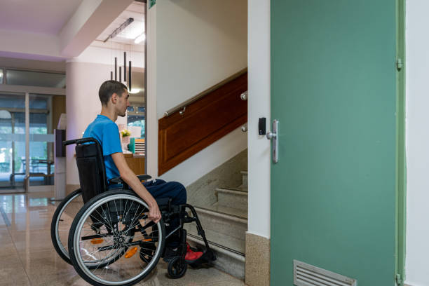 A disabled teenager with dystrophy in a blue T-shirt in a wheelchair stands in front of an obstacle, in front of the stairs. stock photo