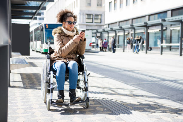 disabled woman in a wheelchair waiting at a bus station and using smart phone - wheelchair street imagens e fotografias de stock