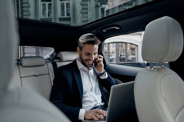 Discussing business details. Handsome young man in full suit talking on smart phone and smiling while sitting in the car back seat stock pictures, royalty-free photos & images
