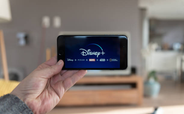 迪士尼®手機啟動畫面。迪士尼+線上視頻，內容流訂閱服務。男人舉起他的智慧手機，看著迪士尼加 - disney 個照片及圖片檔