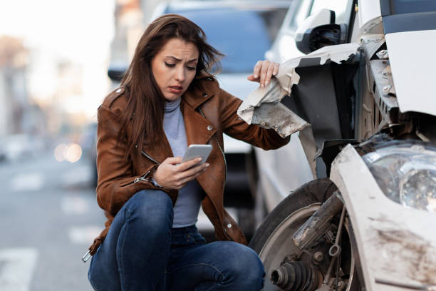 Displeased woman dialing for help after a car accident in the city. Young sad woman text messaging on smart for after a car crash on the road. accidents and disasters photos stock pictures, royalty-free photos & images