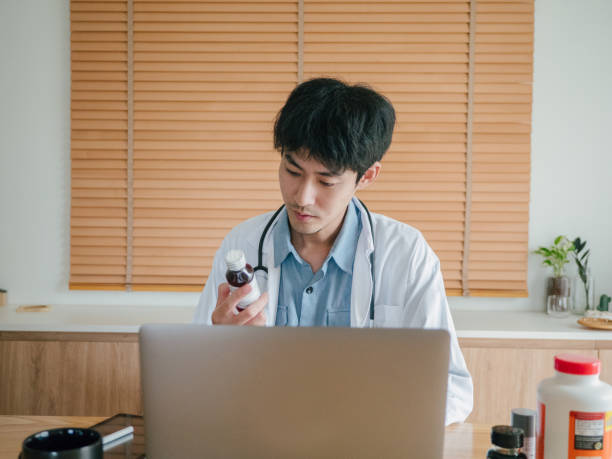 Distance Medical Help. Asian man medical worker sitting at laptop at home, making notes in patient card and talking via internet to patient. doctor of physiotherapy online stock pictures, royalty-free photos & images