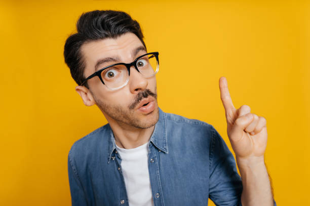Distorted portrait of amazed surprised caucasian guy in a glasses and in an orange t-shirt, shocked looking at the camera, shows IDEA gesture, standing against an isolated orange background stock photo