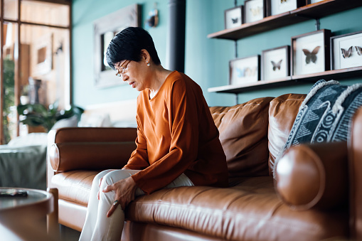 Una mujer asiática mayor angustiada que se siente mal, sufre de dolor en la pierna mientras está sentada en el sofá de la sala de estar de su casa photo